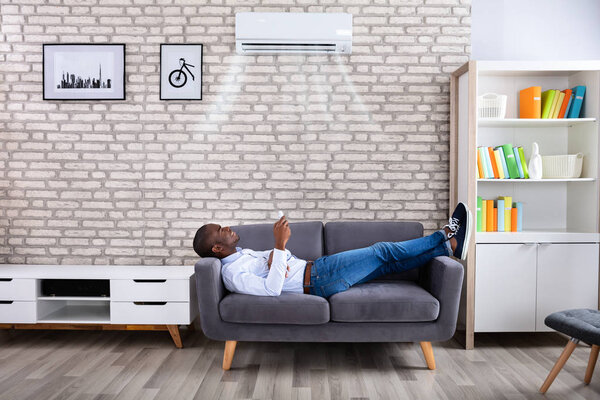 Young African Man Relaxing On Sofa Operating Air Conditioner With Remote Control