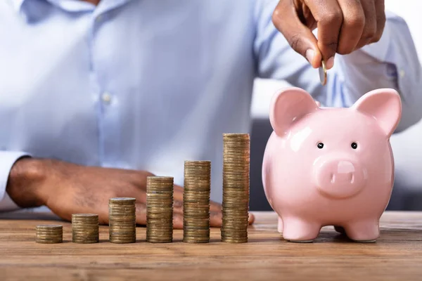 Africano Hombre Insertando Moneda Piggybank Cerca Pila Monedas — Foto de Stock