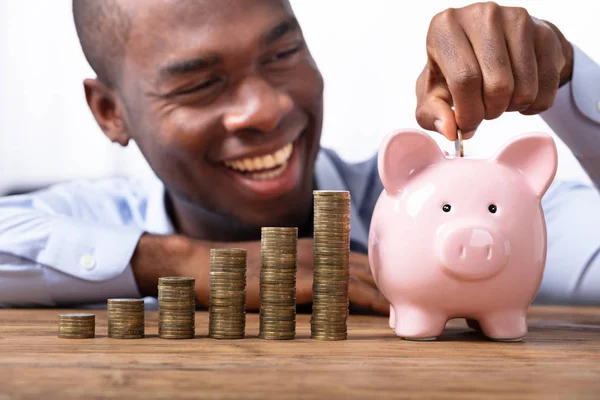 Hombre Africano Feliz Insertando Moneda Piggybank Cerca Pila Monedas —  Fotos de Stock
