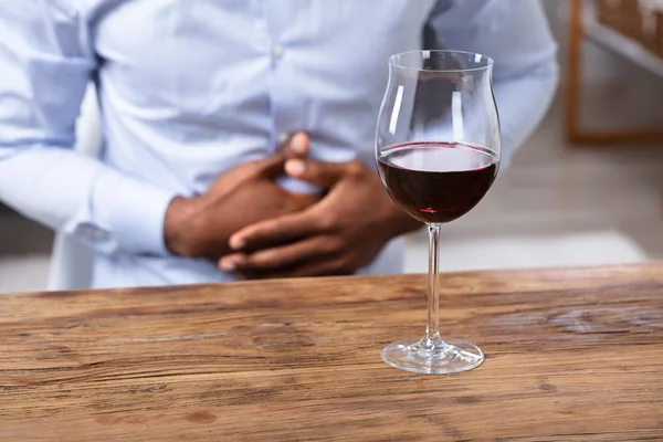 Glass Of Wine On Wooden Desk In Front Of Man Holding His Stomach