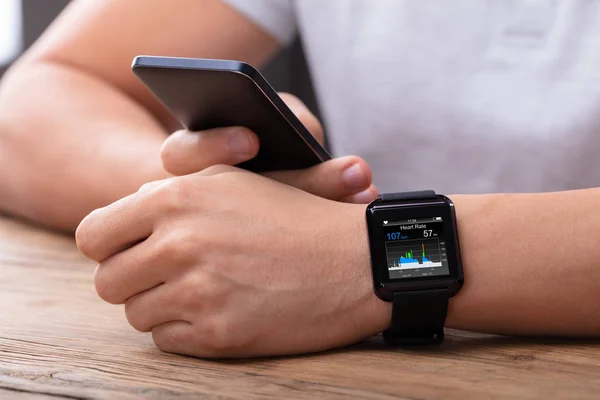 Männerhand Mit Handy Und Smartwatch Zeigt Herzfrequenz Über Holztisch — Stockfoto