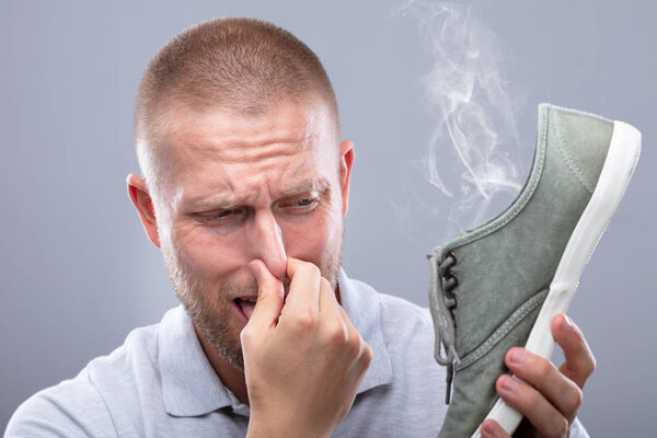 Close-up Of A Man Covering His Nose While Holding Stinky Shoe On Grey Background