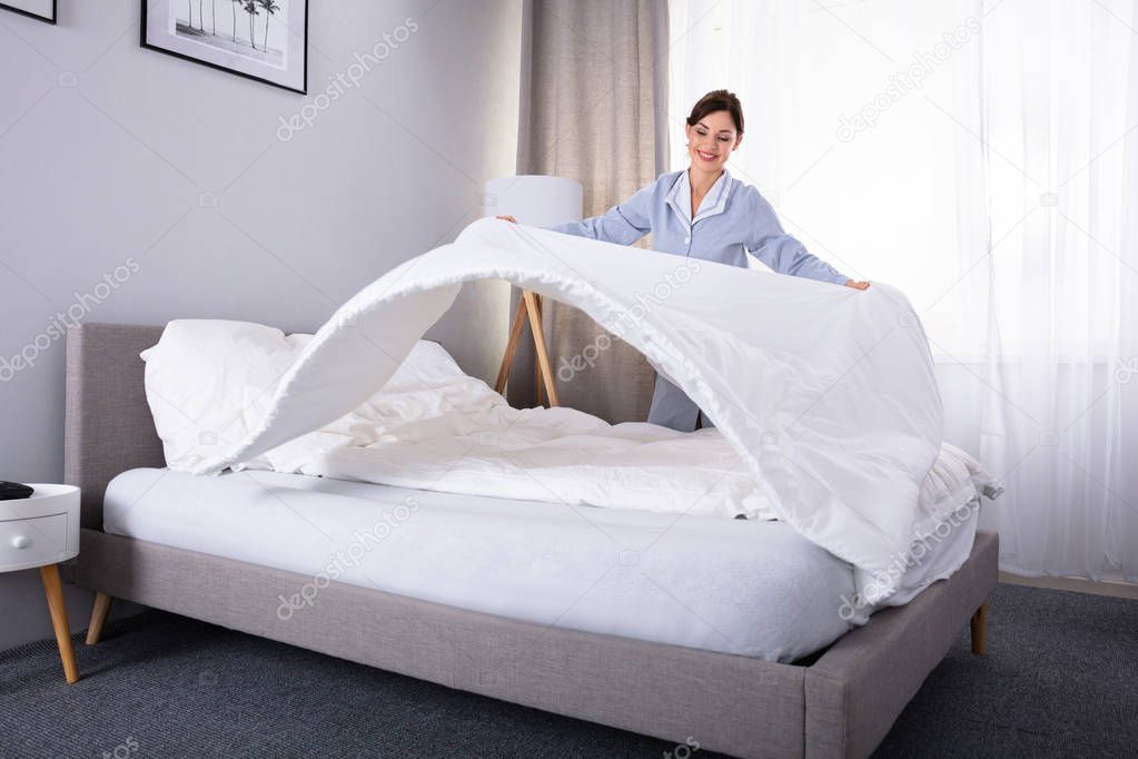 Smiling Young Female Housekeeper Arranging Bedsheet On Bed In Hotel Room