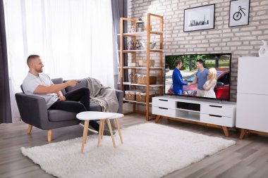 Oturan adam evde televizyon seyrediyor.