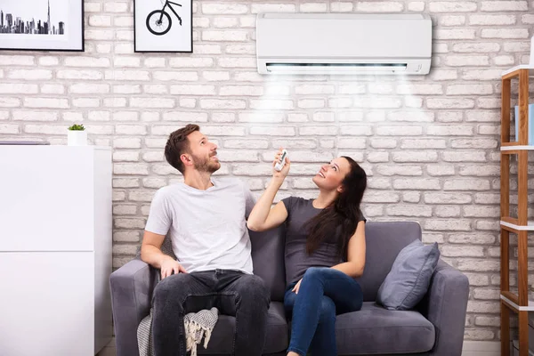 Happy Young Couple Sitting Sofa Operating Air Conditioner Home — Stok Foto