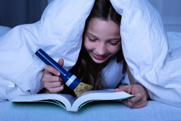 在白色毯子下的女孩使用手电筒 而阅读书籍在夜间 — 图库照片