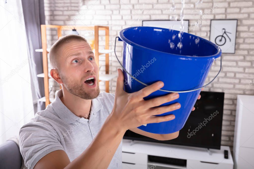 Close-up Of A Young Man Holding Bucket While Water Droplets Leak From Ceiling At Home