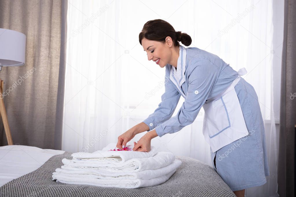 Side View Of A Happy Young Housemaid Placing Flowers On Stack Of Towels Over Bed