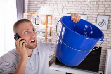 Young Man Collecting Water Leakage In Bucket While Calling Plumber On Smartphone clipart