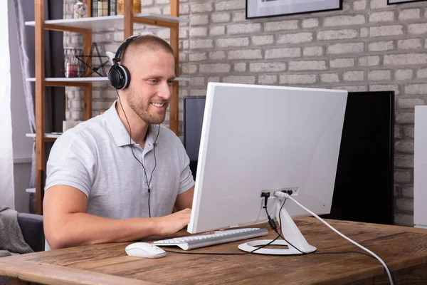 木製の机の上でコンピュータに取り組んでいる間 ヘッドフォンを使用して笑顔若い男 — ストック写真