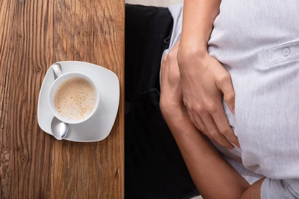 在木制桌子上端着一杯咖啡从胃痛中痛苦的人 — 图库照片