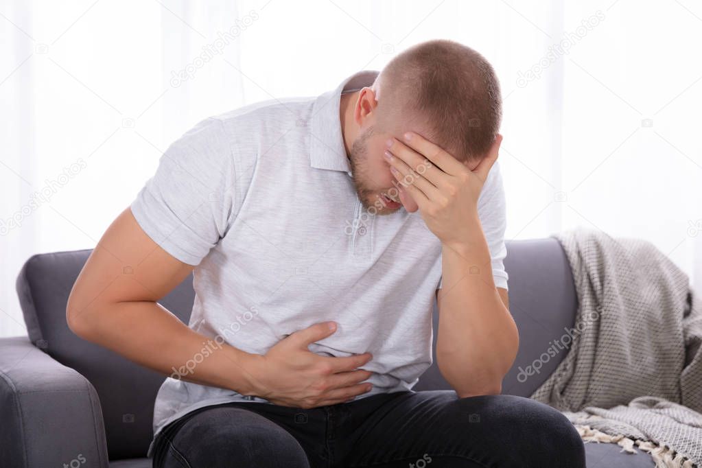 Sad Young Man Sitting On Sofa Suffering From Stomach Pain