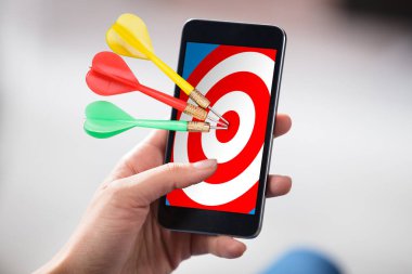 Close-up Of A Person's Hand Holding Cellphone With Colorful Darts On Target clipart