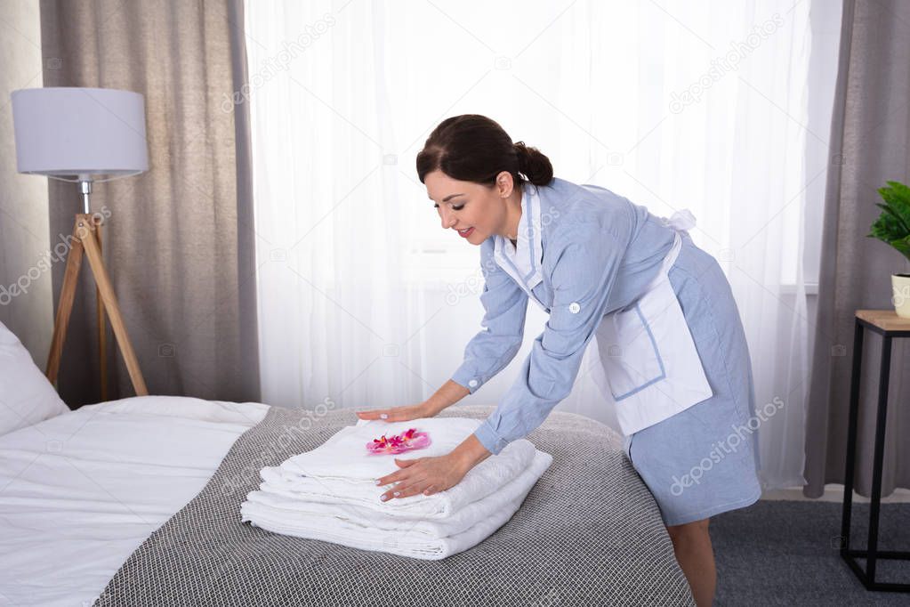 Side View Of A Happy Young Housemaid Placing Flowers On Stack Of Towels Over Bed