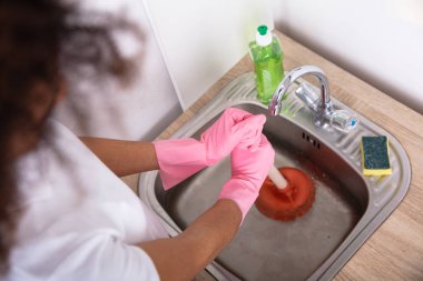 Close-up Of A Female Plumber Wearing Pink Gloves Using Plunger In The Kitchen Sink clipart