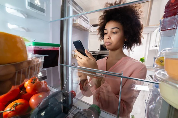 携帯電話を使用して料理を冷蔵庫の前に立って幸せな若い女 — ストック写真