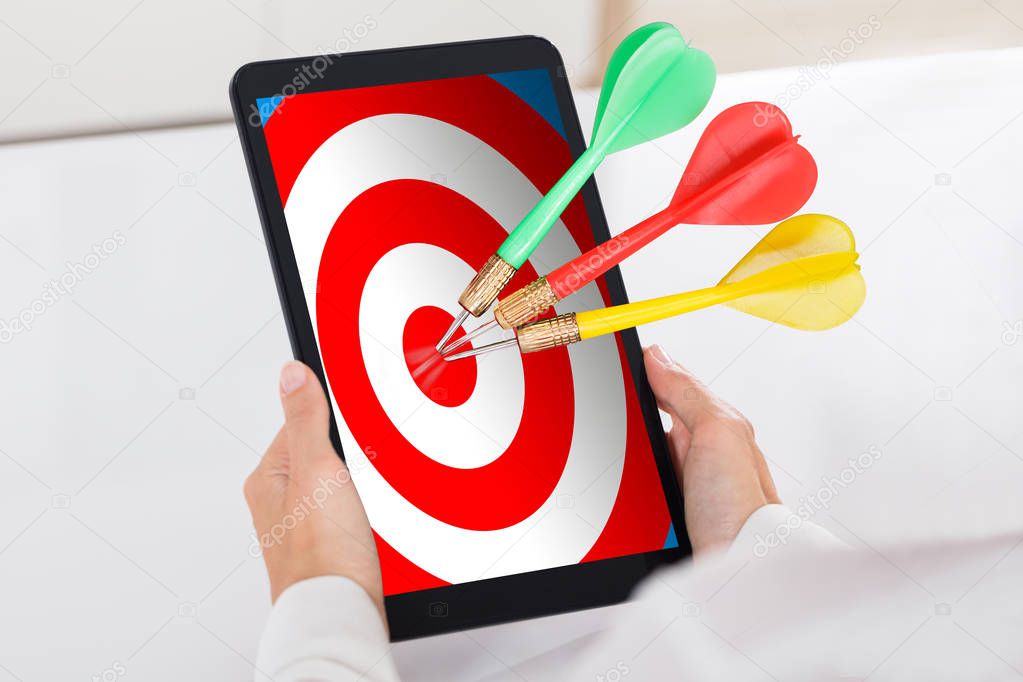 Human Hand Holding Digital Tablet With Colorful Darts On Target Over White Desk