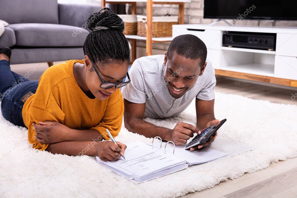 Young African Couple Lying On Carpet Invoice With Calculator