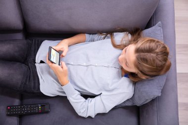 Cep telefonundan kısa mesaj yazarak kanepe üzerinde rahatlatıcı bir kızın yüksek açılı görünüş