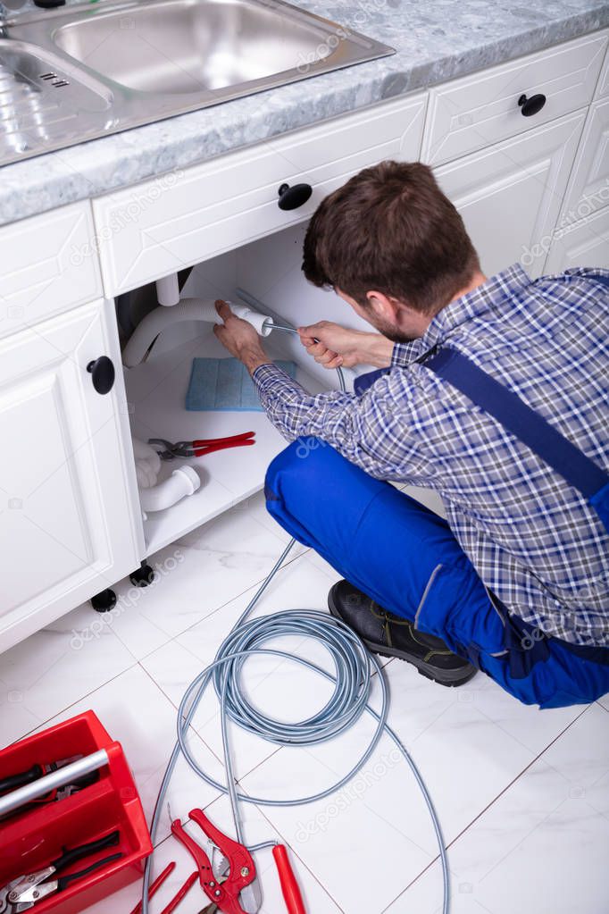 Young Male Plumber Cleaning Clogged Sink Pipe In Kitchen
