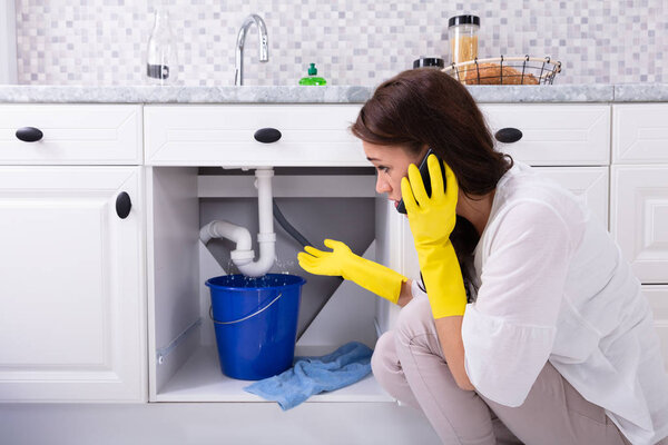 Sad Young Woman Calling Plumber In Front Of Water Leaking From Sink Pipe