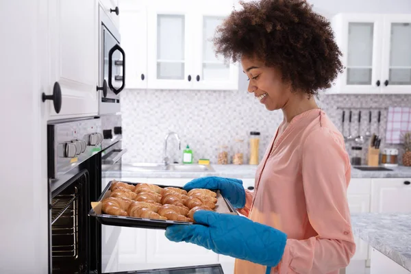 一个快乐的年轻女子穿着蓝色的米特从烤箱上去除烤羊角面包托盘 — 图库照片
