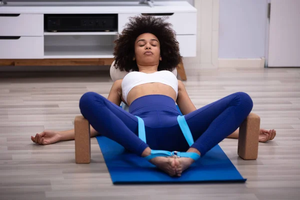 Relaxed Young Woman Lying On Fitness Mat Doing Exercise With Yoga Belt And Two Blocks