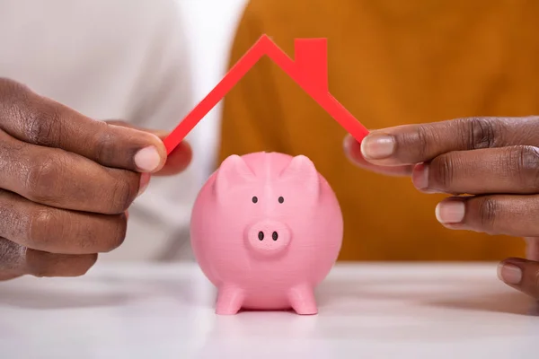 Mão Humana Protegendo Piggybank Rosa Com Telhado Vermelho Sobre Mesa — Fotografia de Stock