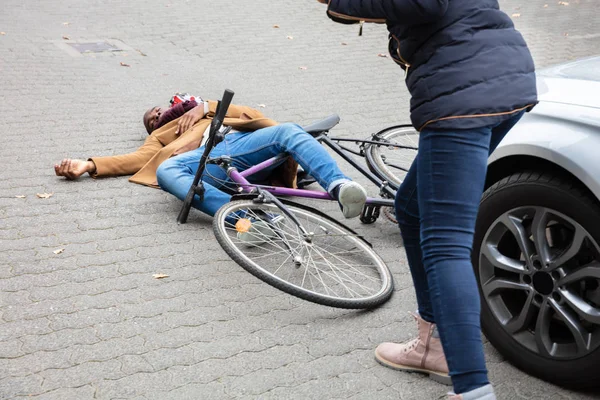 年轻女子看着无意识的男子自行车运动员躺在汽车附近的大街上 — 图库照片