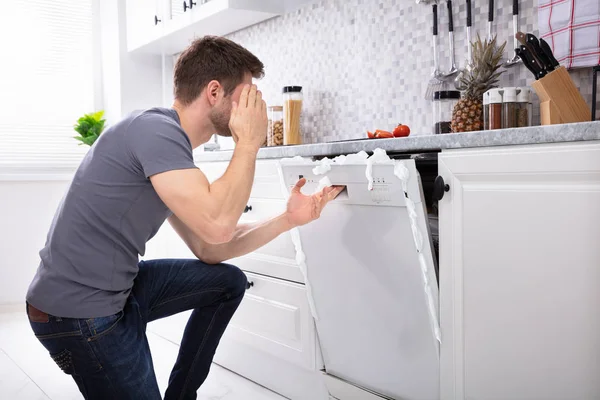 Upset Man Sitting In Front Of Damaged Dishwasher With Foam Coming From It