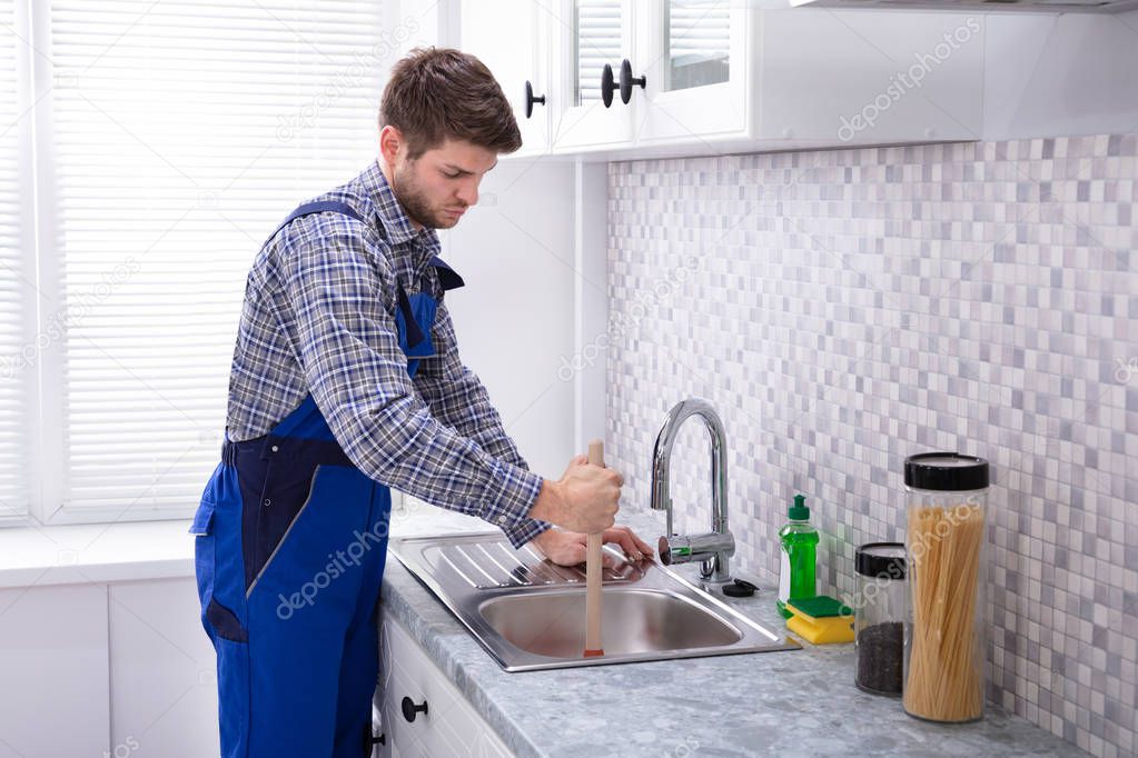 Side View Of A Male Plumber Using Plunger In Kitchen Sink