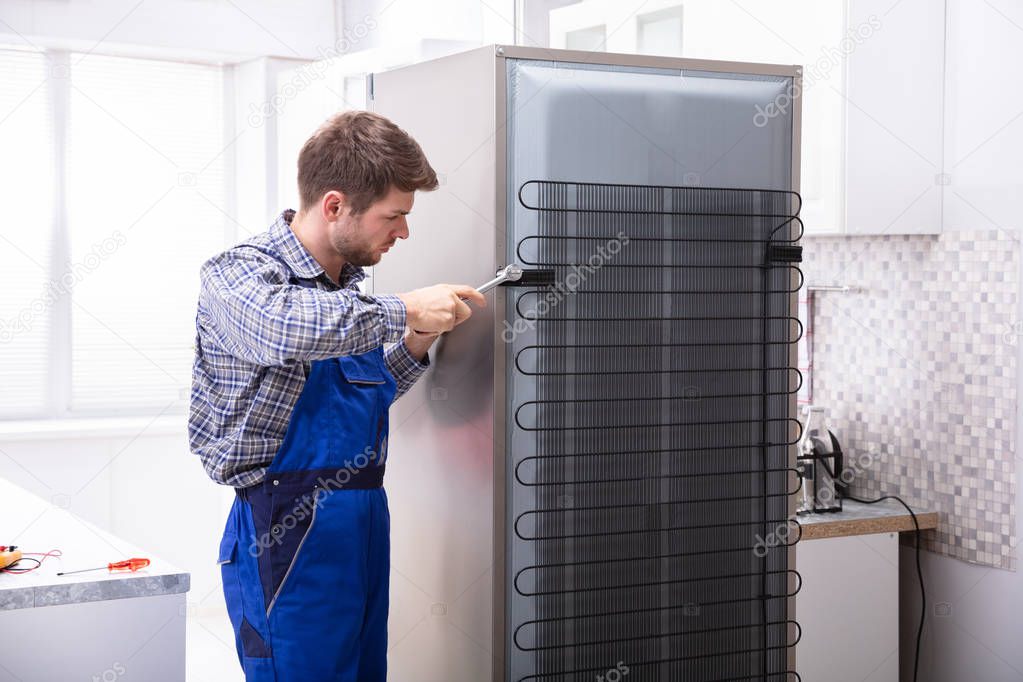 Serviceman In Overall Working On Fridge With Wrench In Kitchen