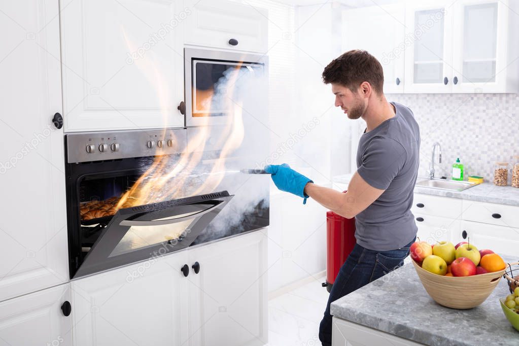 Man Using Fire Extinguisher To Stop Fire Coming Out From Oven In Kitchen