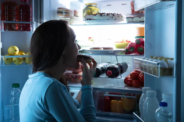 一个年轻女子站在冰箱前吃甜甜圈的侧视图 — 图库照片