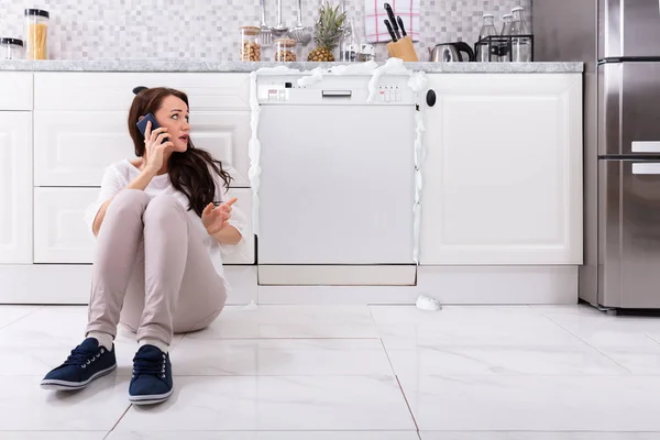 Sad Woman Calling Technician On Cellphone To Fix Dishwasher With Foam Coming From It