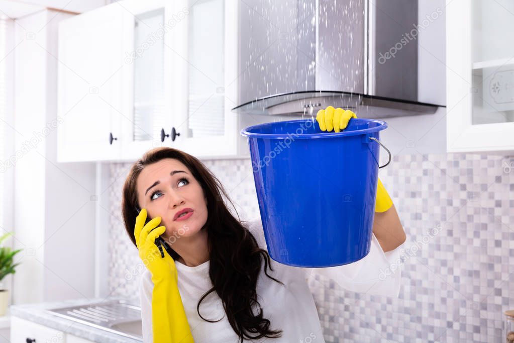 Worried Young Woman Calling Plumber On Cellphone And Collecting Water Leaking From Ceiling In Bucket