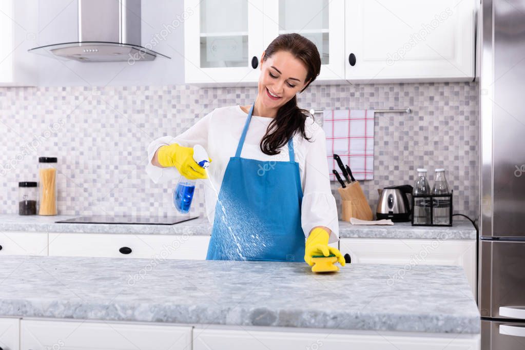 Smiling Female Janitor In Uniform Cleaning Kitchen Counter With Spray Bottle