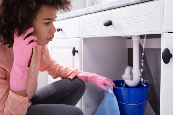 Young Woman Wearing Pink Gloves In Hand Calling Plumber In Front Of Leaking Sink Pipe