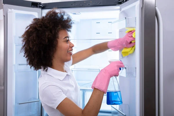 年轻快乐清洁女士清洁空冰箱门与喷雾瓶和餐巾 — 图库照片