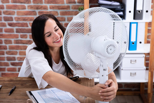 Улыбающаяся молодая женщина наслаждается бризом с электрическим вентилятором в офисе
