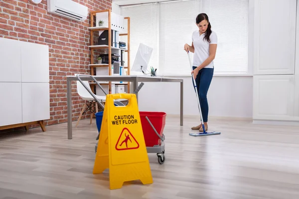 用拖把擦拭靠近警告湿地板标志的妇女清洁办公室 — 图库照片