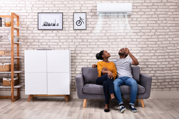 Young African Couple Sitting On Sofa Operating Air Conditioner At Home