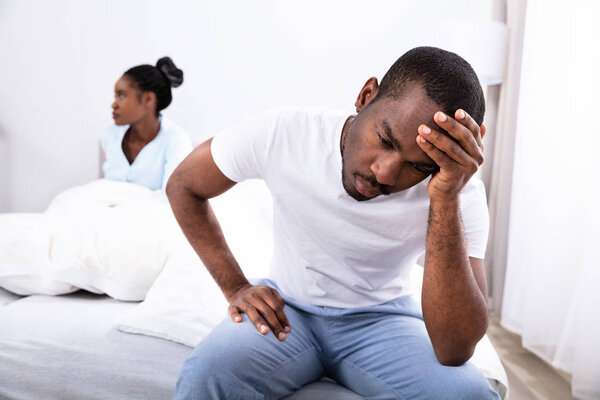 Расстроенный молодой африканский мужчина, сидящий на кровати перед своей женой
