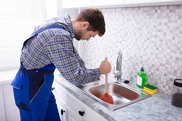 Side View Of A Male Plumber Using Plunger In Kitchen Sink