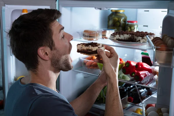 オープン冷蔵庫の近くでケーキを食べる幸せな男の肖像画 — ストック写真