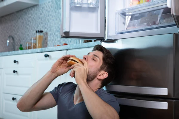 Närbild Man Som Äter Hamburgare Lutar Sig Mot Kylskåp — Stockfoto