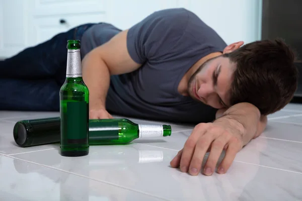 酒精中毒的男子躺在厨房的地板上 手里拿着啤酒瓶 — 图库照片