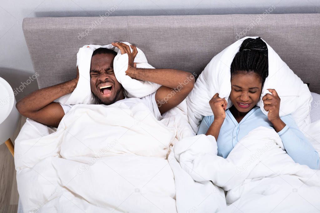 Young African Couple Disturbed By Noise Covering Their Ears With Pillow On Bed