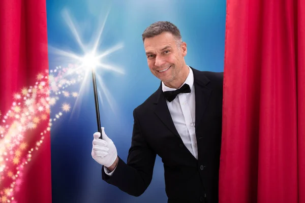 赤いカーテンの後ろに魔法の杖を保持する幸せな成熟した男性魔術師 — ストック写真