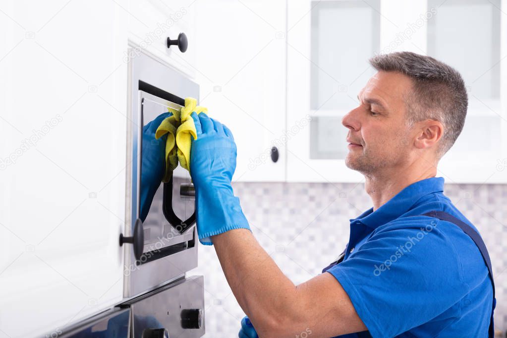 Side View Of Male Janitor Cleaning Oven With Yellow Napkin In Kitchen